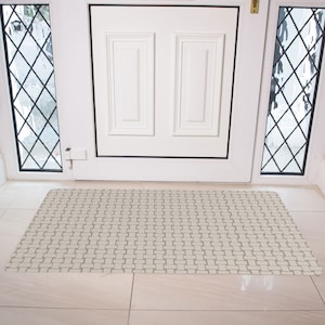 May include: A white doormat with a woven pattern in a light gray color. The doormat is placed in front of a white door with a black trim.