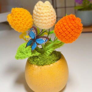 Pode incluir: Um arranjo de flores em crochê com três flores arredondadas em amarelo, creme e laranja, acompanhadas por folhas verdes de crochê e um ornamento de borboleta azul, em um vaso de cerâmica amarelo em forma de ovo com musgo verde.