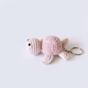 May include: A light pink crocheted turtle keychain featuring a textured shell, small flippers, and two black bead eyes. The soft yarn creature is attached to a circular silver-toned metal split ring, providing a functional way to secure keys.