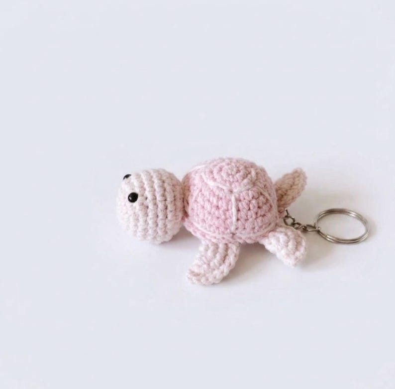 May include: A light pink crocheted turtle keychain featuring a textured shell, small flippers, and two black bead eyes. The soft yarn creature is attached to a circular silver-toned metal split ring, providing a functional way to secure keys.