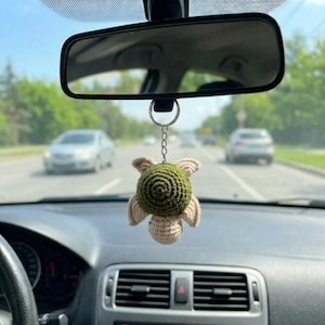 May include: A crochet turtle hanging ornament featuring a dark olive green shell with a spiral pattern and light cream-colored limbs and head. The small stuffed animal is attached to a silver-toned metal split ring for hanging from a rearview mirror.