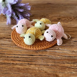 May include: A collection of four small crocheted turtle keychains in pastel pink, light green, and yellow colors. Each turtle features a round shell, a small head with black bead eyes, and four flippers. One pink turtle has a metal keyring attached.