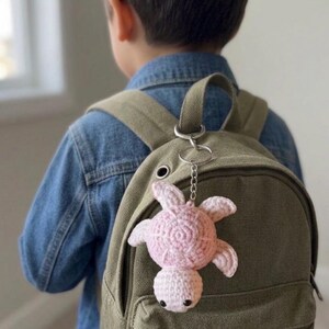 May include: A light pink crocheted sea turtle keychain attached to a solid olive green canvas backpack. The turtle features a round shell with a textured spiral pattern, four small flippers, and a small head with two black bead eyes.