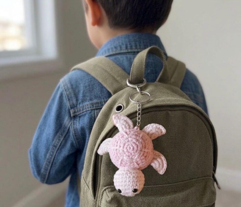 May include: A light pink crocheted sea turtle keychain attached to a solid olive green canvas backpack. The turtle features a round shell with a textured spiral pattern, four small flippers, and a small head with two black bead eyes.