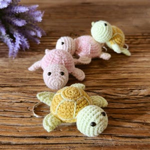 May include: Three crocheted turtle keychains featuring small black bead eyes. One turtle is yellow and light green, while the other two are light pink. Each turtle has a metal keyring attached to its body, showcasing detailed yarn stitching.