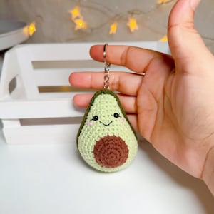 Könnte beinhalten: Ein gehäkelter Avocado-Schlüsselanhänger mit einem hellgrünen, strukturierten Körper, einem dunkelgrünen Rand und einem braunen Kern in der Mitte. Die Avocado hat gestickte schwarze Augen, ein kleines schwarzes Lächeln und zwei winzige rosa Wangen.