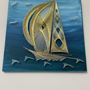 Könnte beinhalten: Ein handgefertigtes String-Art-Stück, das ein Segelboot auf einem blau bemalten Hintergrund darstellt. Das Segelboot besteht aus goldenem Faden und kleinen Nägeln, wodurch ein strukturierter Effekt entsteht. Das Kunstwerk enthält kleine, weiße Nagelakzente.