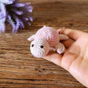 May include: A small, light pink crocheted turtle keychain featuring a textured shell, a rounded head with two black bead eyes, and four short flippers. The turtle is crafted from yarn and includes a metal ring attachment for keys.