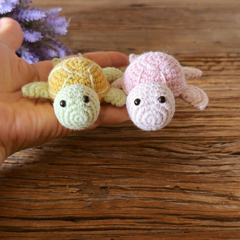 May include: Two small crocheted turtle toys. One turtle has a light green head and a yellow shell with white stitching. The other turtle is entirely light pink. Both have round black plastic eyes and four small flippers.