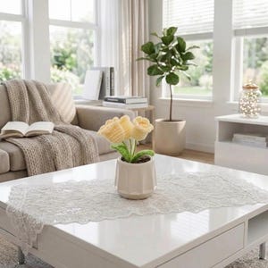 May include: A crochet flower arrangement featuring three pale yellow tulip-shaped blossoms with green stems and leaves, potted in a small, smooth, off-white ceramic container with a subtle vertical ribbed texture on the exterior surface.