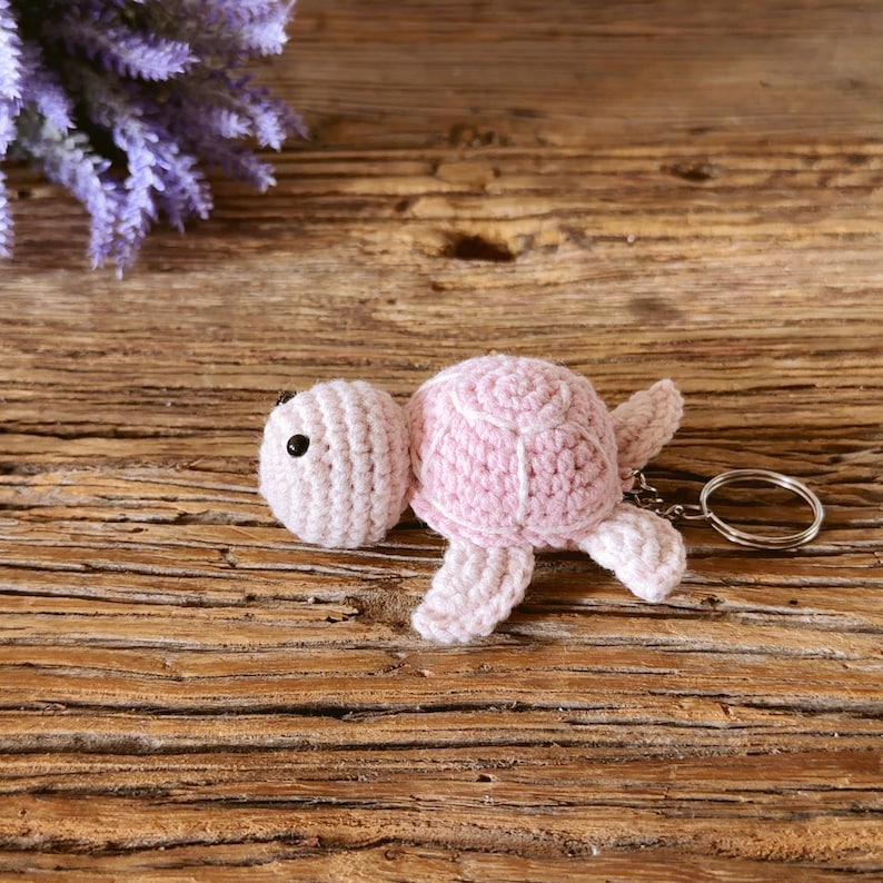 May include: A light pink crocheted turtle keychain featuring a rounded shell, four small flippers, and a small black bead for an eye. A silver-toned metal split ring is attached to the back of the turtle, allowing it to be used as a keychain.