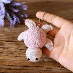 May include: A light pink crocheted sea turtle keychain featuring a round shell with a textured stitch pattern, four small flippers, and a rounded head with two small black bead eyes attached to a silver metal split ring key holder.