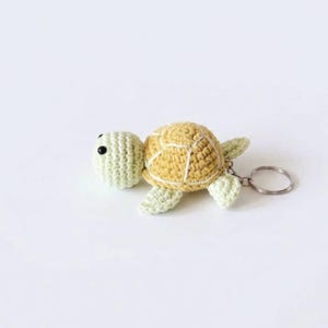 May include: A crochet turtle keychain featuring a pale green head and limbs with a textured yellow shell. The turtle has two small black bead eyes and is attached to a circular silver metal keyring on the right side of its body.