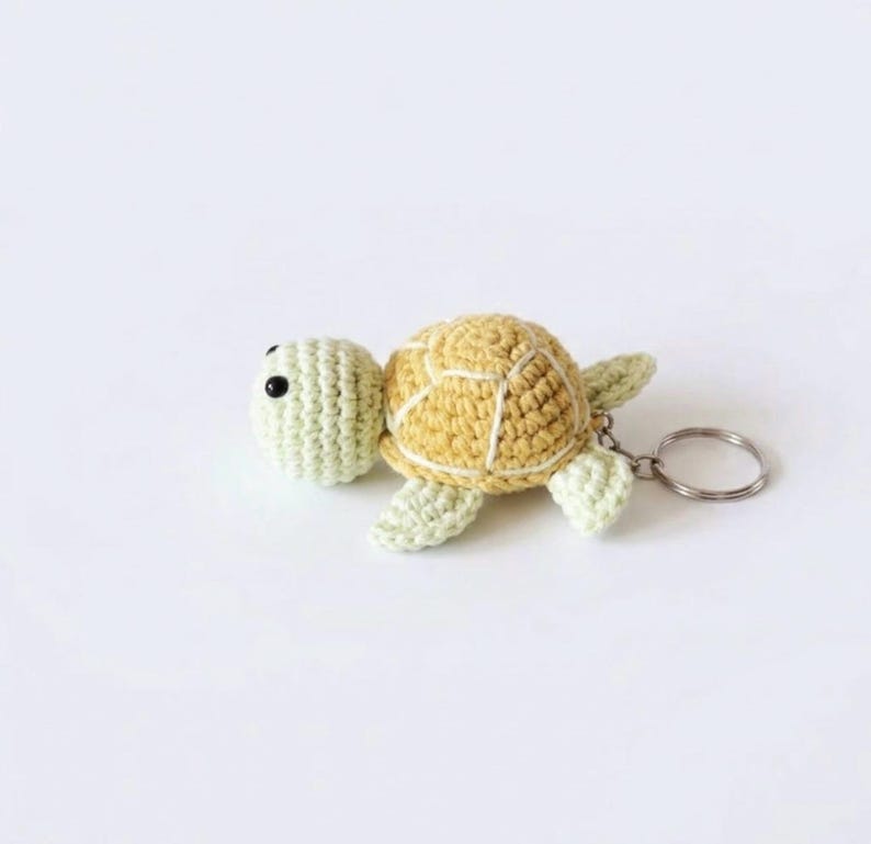 May include: A crochet turtle keychain featuring a pale green head and limbs with a textured yellow shell. The turtle has two small black bead eyes and is attached to a circular silver metal keyring on the right side of its body.