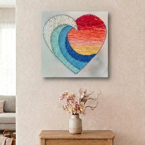 May include: A square string art piece featuring a heart shape composed of layered threads in white, light blue, dark blue, yellow, orange, and red. The design creates a gradient effect with intersecting lines anchored by small metal nails.