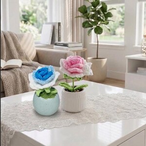 Puede incluir: Dos flores de ganchillo en macetas. Una flor tiene un centro azul brillante con pétalos exteriores blancos en una maceta redonda de color azul claro moteado. La otra flor es rosa con pétalos exteriores blancos, hojas verdes y tallo en una maceta blanca.