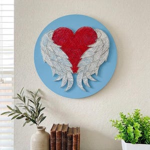 May include: A circular wall art piece featuring string art of a red heart with white wings on a light blue background. The heart and wings are created by winding thread around small nails driven into the circular wooden board surface.