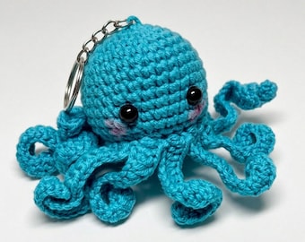 May include: A turquoise crochet octopus keychain featuring a round head with two black plastic eyes and small pink stitched cheeks. The octopus has eight curled tentacles extending outward and a silver metal keyring attached to the top of its head.