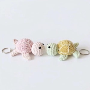 May include: Two crochet turtle keychains with metal rings. One turtle is light pink with black bead eyes, and the other is light yellow and pale green with black bead eyes. Both feature a textured shell pattern and four small flippers.