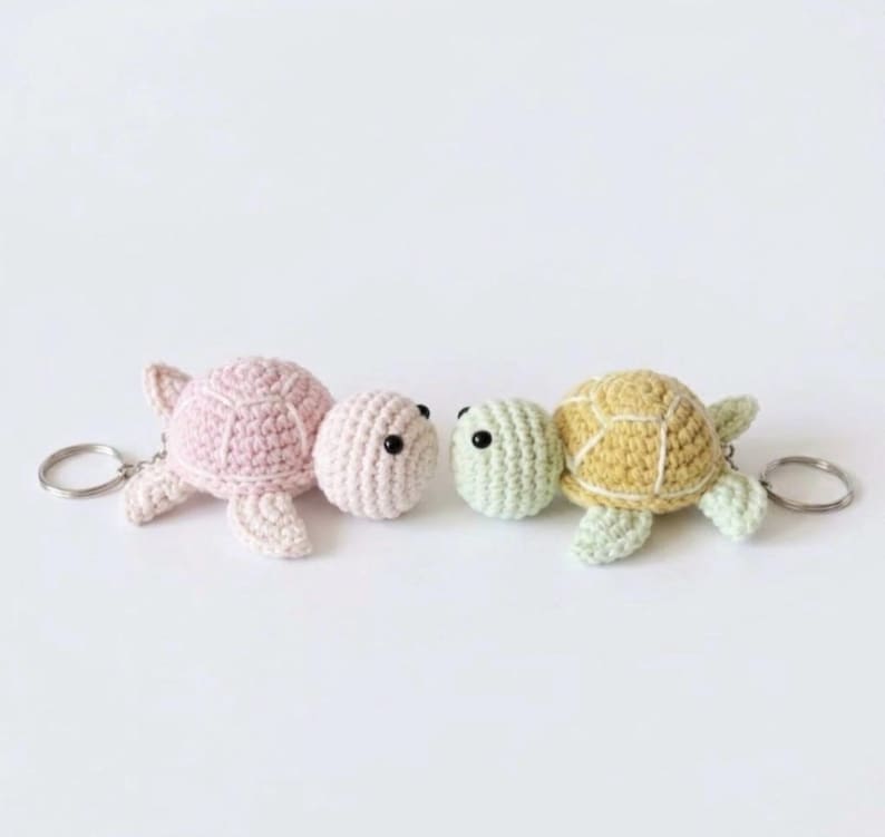 May include: Two crochet turtle keychains with metal rings. One turtle is light pink with black bead eyes, and the other is light yellow and pale green with black bead eyes. Both feature a textured shell pattern and four small flippers.