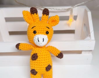 May include: A small, handmade crochet giraffe toy featuring a bright yellow body with dark brown spots, a white muzzle, and dark brown accents on the ears, hands, and feet. The giraffe has black embroidered eyes and small horns on its head.