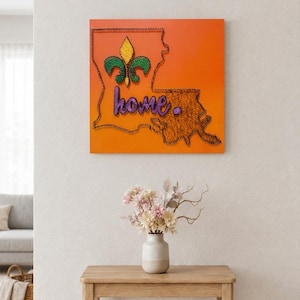 May include: A square orange wall art piece featuring a string art outline of the state of Louisiana. Inside the outline is a yellow and green fleur-de-lis and the word home in purple string. The state shape is filled with brown string.