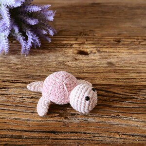 May include: A small, light pink crocheted turtle toy featuring a rounded shell with stitched detailing and two black bead eyes. The soft, handmade yarn creature has four flippers and a textured surface, crafted with a consistent knit pattern.