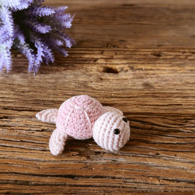 May include: A small, light pink crocheted turtle toy featuring a rounded shell with stitched detailing and two black bead eyes. The soft, handmade yarn creature has four flippers and a textured surface, crafted with a consistent knit pattern.
