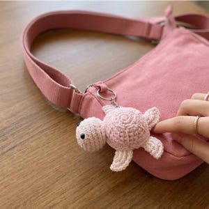 May include: A light pink crocheted turtle keychain attached to a matching pink canvas crossbody bag. The turtle features a rounded shell, four small flippers, and two small black bead eyes, secured by a silver-toned metal keyring.