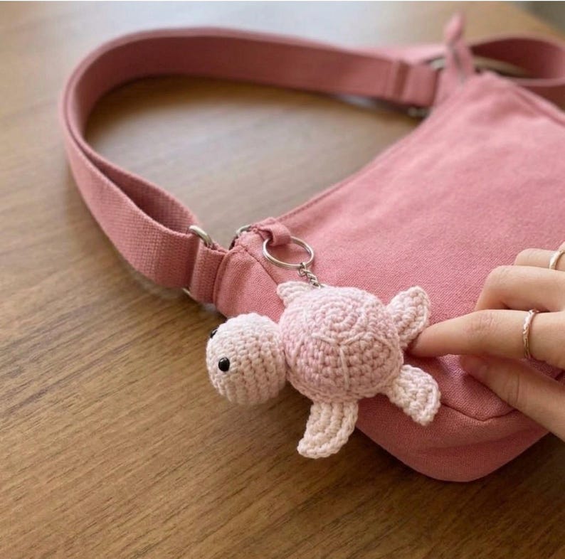 May include: A light pink crocheted turtle keychain attached to a matching pink canvas crossbody bag. The turtle features a rounded shell, four small flippers, and two small black bead eyes, secured by a silver-toned metal keyring.