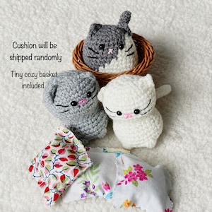 May include: Three small crocheted cat figures in gray, white, and mixed gray and white colors. One cat sits inside a round woven brown basket. Two small fabric cushions with floral patterns are placed in front of the cat figures.