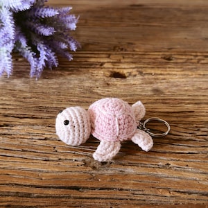 May include: A light pink crocheted turtle keychain featuring a small round head with a black bead eye and a textured shell. The soft yarn creature is attached to a circular silver-toned metal split ring for keys.