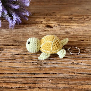May include: A crochet turtle keychain featuring a pale green head and limbs with a textured yellow shell. The turtle has a small black bead eye and is attached to a circular silver-toned metal split ring for keys or bag attachment.