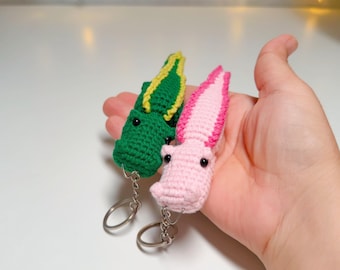 May include: Two small crochet keychains shaped like axolotls, one in vibrant green with yellow accents and the other in soft pink with darker pink details. Each creature features black bead eyes and a metal split ring attached for keys.