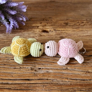 May include: Two crocheted turtle keychains. One turtle has a mustard yellow shell with a light green head and flippers. The second turtle is light pink with a matching head and flippers. Both have small black bead eyes and a metal keyring.