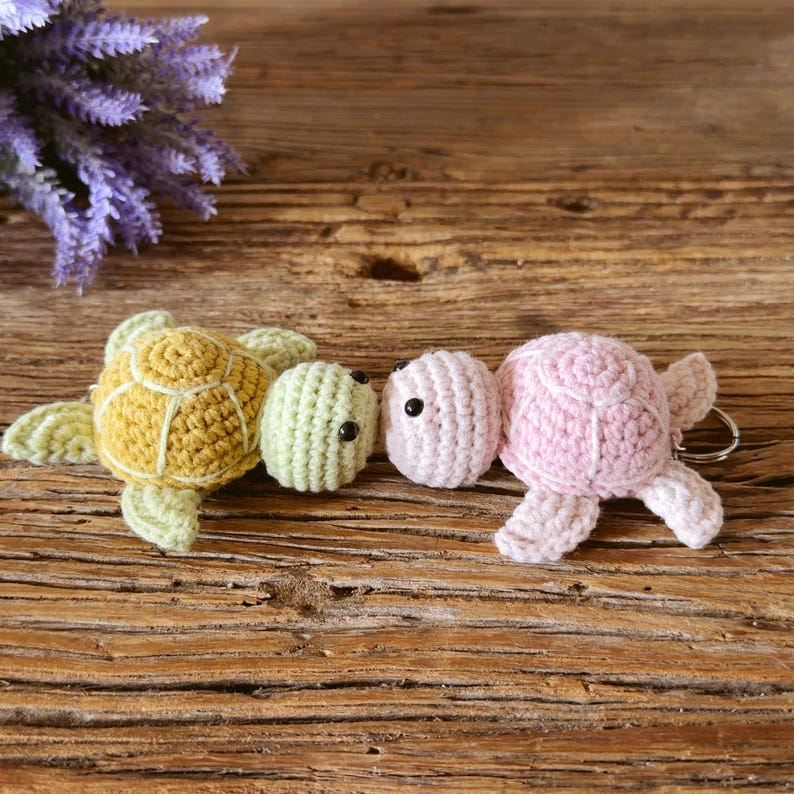 May include: Two crocheted turtle keychains. One turtle has a mustard yellow shell with a light green head and flippers. The second turtle is light pink with a matching head and flippers. Both have small black bead eyes and a metal keyring.