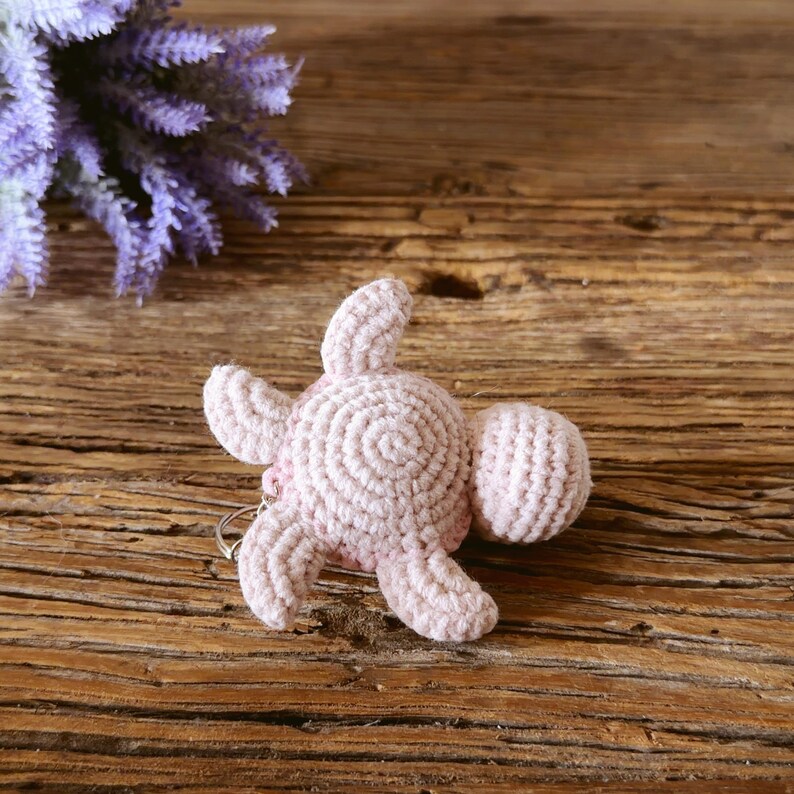 May include: A light pink crochet sea turtle keychain featuring a textured shell with a spiral pattern, four flippers, and a small head, attached to a silver metal ring. The turtle is crafted from yarn with visible stitches.