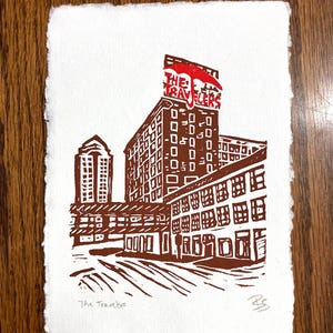 May include: A brown and red linocut print on white paper featuring a city building with a red umbrella icon and the text THE TRAVELERS on the roof. The artwork depicts a multi-story structure with many windows and the handwritten title The Travelers.