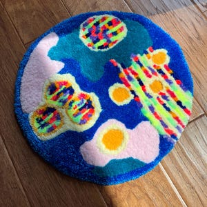 May include: A circular tufted rug featuring a vibrant blue background with abstract shapes in neon yellow, pink, teal, and orange. The design includes textured patterns of multicolored stripes, solid circles, and organic blobs with a plush pile finish.