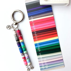 May include: A keychain featuring two strands of multi-colored clay disc beads with silver spacers, a small yin-yang charm, and a silver lobster clasp, positioned next to a color palette card labeled clear bright vivid icy and high contrast value.