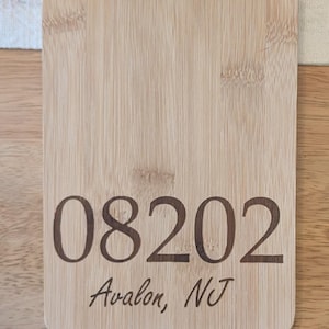 May include: A light-colored bamboo cutting board with a handle and a hole for hanging. The numbers "08202" and the text "Avalon, NJ" are etched into the wood. The board is rectangular with rounded corners.