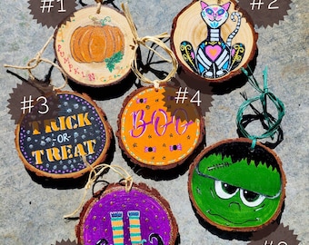 May include: Hand-painted Halloween ornaments on wood slices. Designs include a pumpkin, a cat, the words "Trick or Treat", the word "Boo", witch legs, and Frankenstein's monster. Each ornament has a twine loop for hanging.