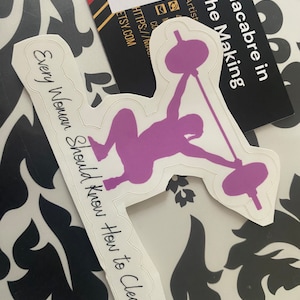 May include: A white sticker with a purple silhouette of a person lifting a barbell. The text "Every Woman Should Know How to Clean" is written in a cursive font on the sticker.