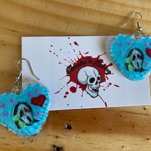 Könnte beinhalten: Ein Paar blaue herzförmige Ohrringe mit einem weißen und grünen Bild einer maskierten Figur darin. Die Ohrringe hängen an silbernen Haken. Der Text "Love Me Dead" ist auf dem Herzen sichtbar.