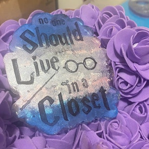 Pode incluir: Um ornamento de resina azul e prata com o texto "No one should live in a closet" e um par de óculos. O ornamento é rodeado por rosas de espuma roxa.