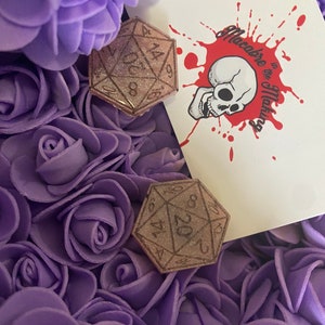 May include: Two brown resin dice with glitter embedded in the resin. The dice are 20-sided and have the numbers 1 through 20 printed on them. The dice are sitting on a bed of purple artificial flowers. The background is a white card with a red skull and the text "Macabre to the Maxing".