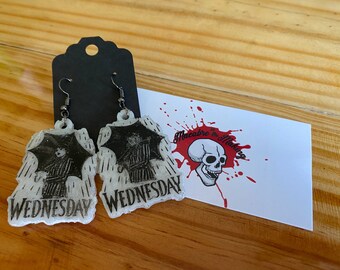 Może przedstawiać: Para białych, błyszczących kolczyków z czarno-białym obrazkiem Wednesday Addams trzymającej parasol w deszczu. Kolczyki są oznaczone napisem "WEDNESDAY" i wiszą na srebrnych haczykach. Kolczyki są prezentowane na drewnianej powierzchni z wizytówką, na której widnieje napis "Macabre in the Making" i grafika czaszki.