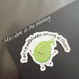 May include: A sticker featuring a cartoon gallbladder character with arms and legs, smiling. The sticker reads "I got my gallbladder removed at Claire's." Above the sticker, the text "Macabre in the Making" is written.