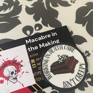 Pode incluir: Um autocolante redondo com o texto "WORKING IN DEATH CARE ISN'T CREEPY" a rodear um caixão castanho com flores brancas. Um cartão de visita com uma caveira e as palavras "Macabre in the Making" também é visível.