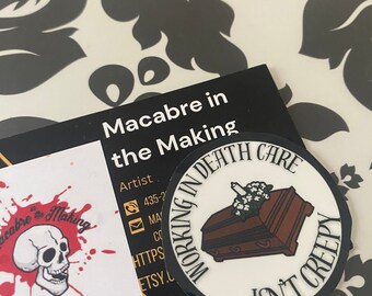 May include: A circular sticker featuring the text "WORKING IN DEATH CARE ISN'T CREEPY" around a brown coffin with white flowers. A business card with a skull and the words "Macabre in the Making" is also visible.